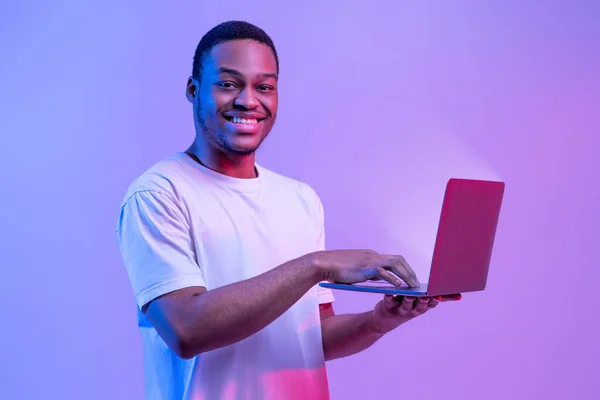 Online Work. Portrait Of Smiling African Guy Using Laptop With Glowing Screen