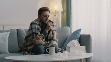 Ekoseye sarılı, öksürük krizinden muzdarip, iyileştirici çay içen, evdeki kanepede tek başına oturan genç hasta adam.