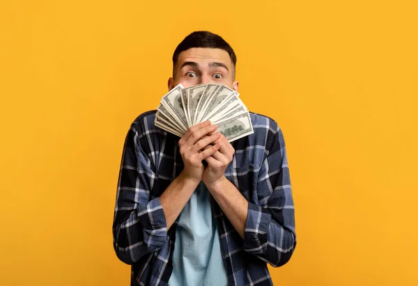Emocionado millennial chico sosteniendo ventilador de dinero en frente de su cara en naranja fondo del estudio — Foto de Stock