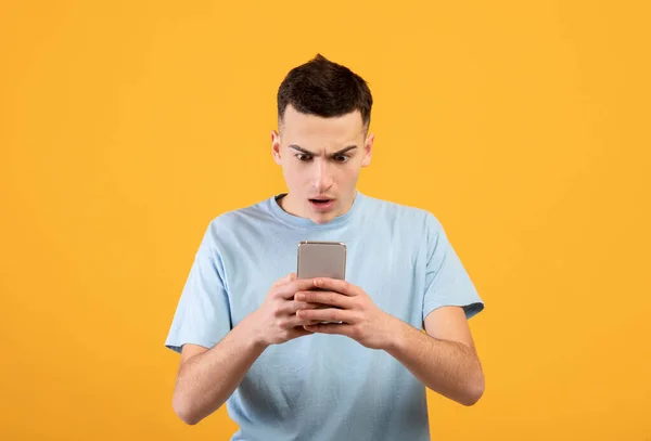 Bonito cara jovem olhando para o telefone móvel em choque sobre fundo estúdio laranja — Fotografia de Stock