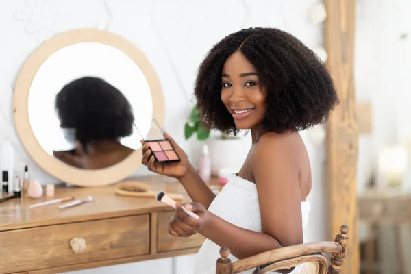 Wanita kulit hitam berdandan di depan cermin, duduk di meja rias dengan palet bayangan mata, tersenyum di depan kamera — Stok Foto