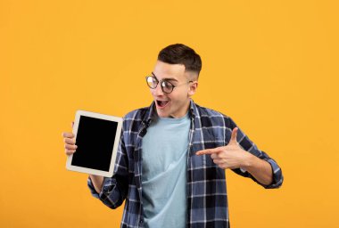 Portakal rengi stüdyo arka planında web sitesi ya da uygulama tasarımı için tablet bilgisayarını gösteren neşeli genç adam