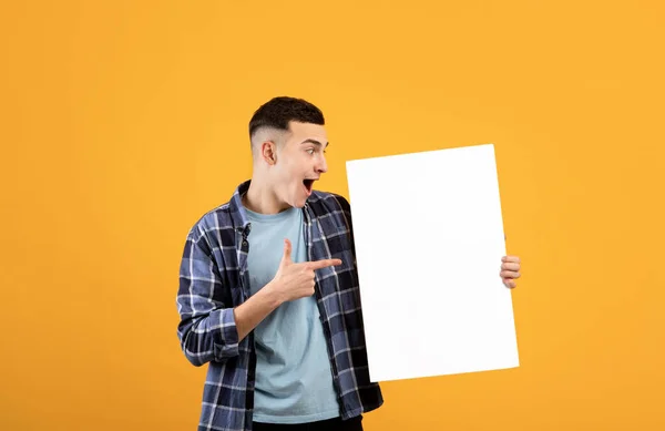 Retrato del joven sorprendido apuntando a un póster de papel en blanco con maqueta para el diseño —  Fotos de Stock