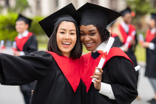 Jovens senhoras alegres em vestes de formatura tomando selfie — Fotografia de Stock