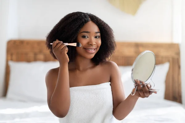 Mulher afro-americana atraente olhando no espelho, aplicando maquiagem na cama em casa. Conceito de rotina diária — Fotografia de Stock