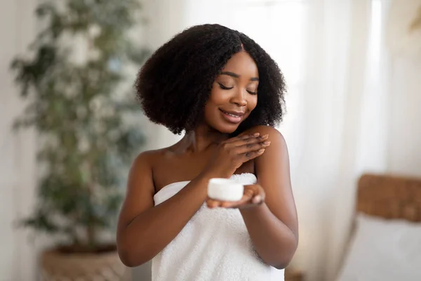 Jeune femme noire enveloppée dans une serviette appliquer lotion pour le corps ou crème après la douche à la maison — Photo