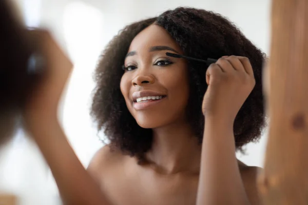 Wanita kulit hitam yang cantik mengenakan maskara di bulu matanya di depan cermin di rumah — Stok Foto