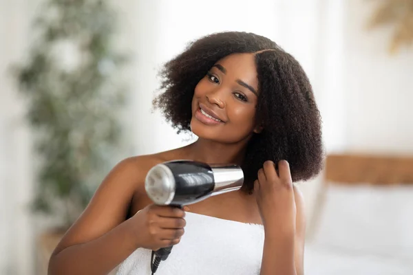 Duş aldıktan sonra saç kurutma makinesi kullanan banyo havlusu içindeki güzel siyah kadın. — Stok fotoğraf