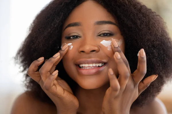 Retrato de una encantadora mujer negra que aplica crema facial en sus mejillas, sonriendo a la cámara, en el interior —  Fotos de Stock