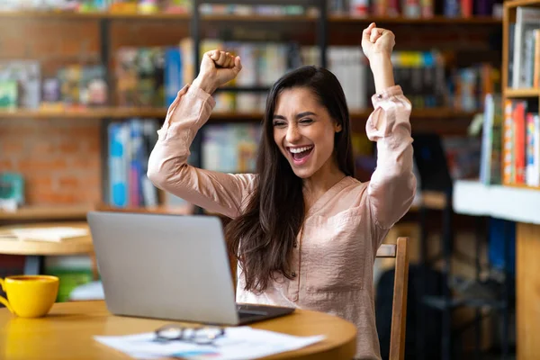 Día de suerte. Mujer latina emocional celebrando el éxito frente a la computadora portátil en la cafetería, apretando puños y gritando — Foto de Stock