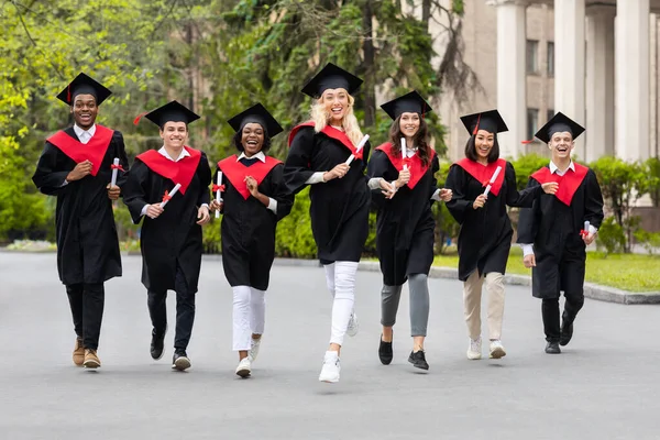 Begeisterte multirassische Studenten in Abschlusskostümen laufen durch den Universitätscampus — Stockfoto