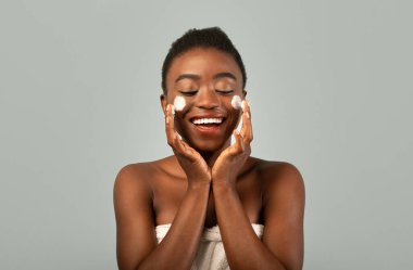 Bakım konsepti. Yanakları kremalı, beyaz havlusu gri arka planda gülümseyen heyecanlı genç siyahi kadın.