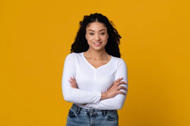 Sarı arka planda katlanmış kollarıyla poz veren kendine güvenen genç zenci bayan.