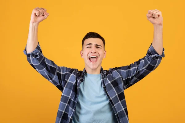 Başarılı genç adam kollarını başının üstüne kaldırıyor, turuncu stüdyo arka planında heyecanla bağırıyor. — Stok fotoğraf