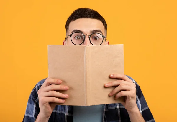 Gözlüklü genç adam kitap arkasına saklanıyor, turuncu arka planda üniversite sınavlarına çalışmakta zorlanıyor. — Stok fotoğraf