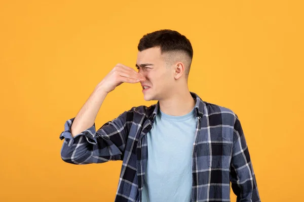 Cool millennial chico cerrando la nariz, sufriendo de olor terrible, no le gusta el mal olor en fondo de estudio naranja —  Fotos de Stock