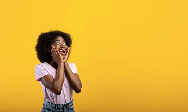 Conceito promocional. Mulher negra incrível olhando para o espaço vazio, tocando seu rosto em gesto surpreso, fundo amarelo — Fotografia de Stock