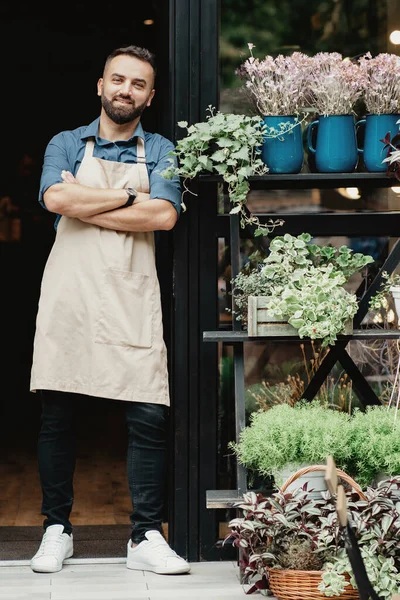 Alegre millennial confiado chico en delantal con brazos cruzados en la puerta principal de la tienda de flores — Foto de Stock