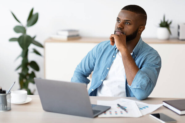 Black Freelancer Guy Sitting At Workplace With Thoughtful Face Expression