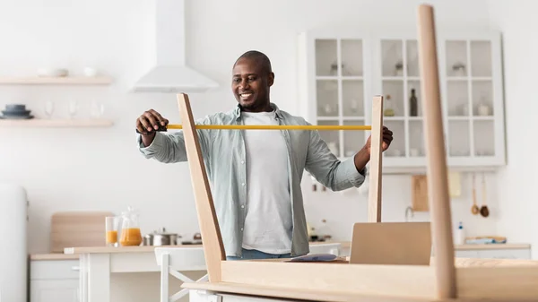 Excited black man measuring furniture with roulette, assembling new table in kitchen interior — Stock Photo, Image