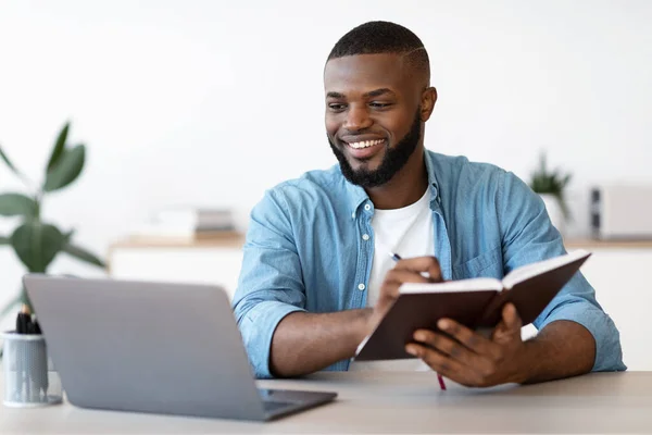 Negro Millennial Freelancer Guy Tomando notas mientras trabaja en el ordenador portátil en casa — Foto de Stock