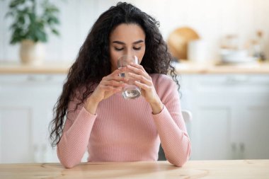 İçecek bir şeyler. Genç kıvırcık esmer kadın mutfaktaki bardaktan maden suyu içiyor.
