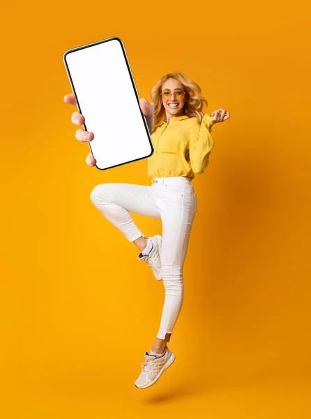 Vidám szőke nő ugrás fel, és mutatja okostelefon üres képernyőn, élvezi az új alkalmazás mobil eszköz — Stock Fotó