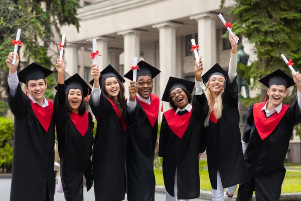 Estudantes multirraciais alegres em trajes de graduação levantando diplomas — Fotografia de Stock