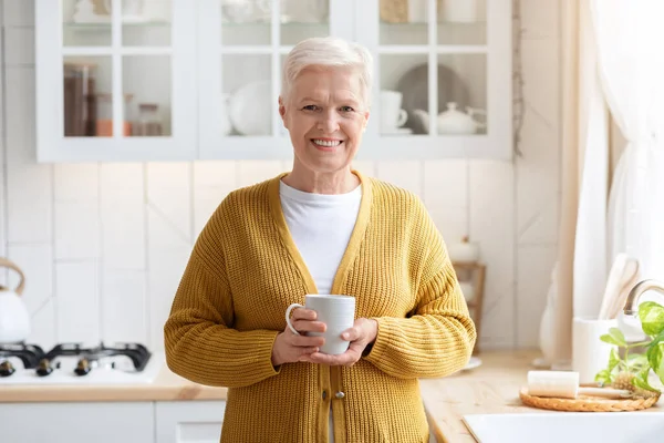 Веселая привлекательная пожилая леди с кружкой, пьет кофе на кухне — стоковое фото