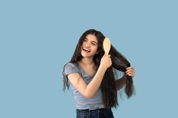 Menina adolescente indiana atraente escovando seu cabelo longo com escova de madeira no fundo do estúdio azul — Fotografia de Stock