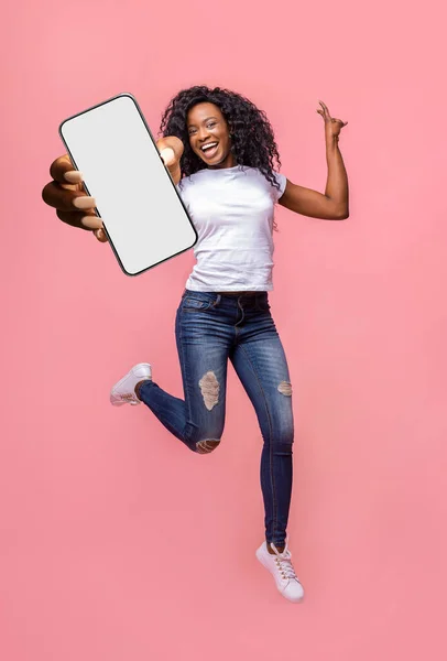 Emocionada mujer negra mostrando smarthone con pantalla blanca en blanco a la cámara, saltando con gadget moderno, maqueta — Foto de Stock