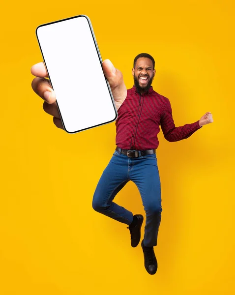 Hombre negro saltando, señalando SÍ y mostrando el teléfono celular con espacio vacío para la aplicación móvil en la pantalla, fondo naranja — Foto de Stock