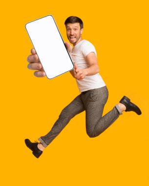 Heyecanlı adam zıplıyor, kamerayı işaret ediyor, turuncu arka planda boş ekranlı akıllı telefonu gösteriyor,