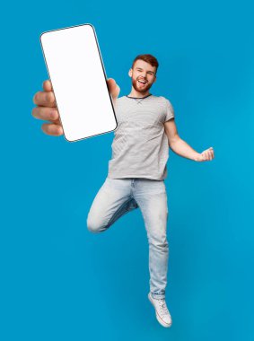 Heyecanlı kızıl adam akıllı telefonuyla zıplıyor, mavi stüdyo arka planında EVET jesti gösteriyor, mobil uygulama için model