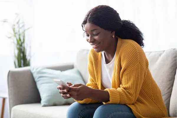 Sorrindo senhora negra sentada no sofá, usando smartphone — Fotografia de Stock
