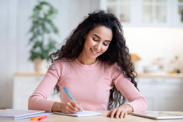 Młoda uśmiechnięta kobieta pisze notatki w notatniku, siedzi przy stole w kuchni — Zdjęcie stockowe