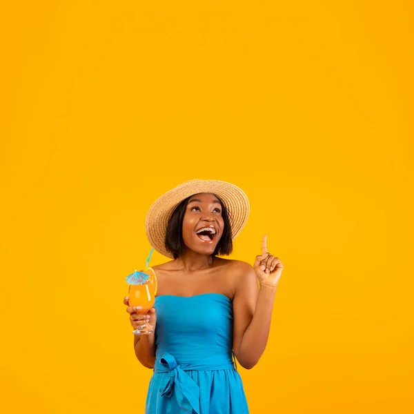 Mulher negra excitada com saboroso coquetel de verão apontando para o espaço vazio acima de sua cabeça, fundo de estúdio laranja — Fotografia de Stock