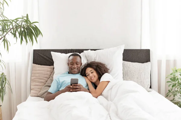 Lächelndes attraktives Millennial-Paar mit Smartphone, gemeinsam im Bett — Stockfoto
