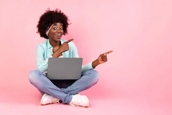 Cheerful Black Woman With Laptop Pointing Fingers Aside, Pink Background