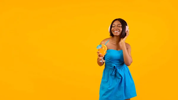 Portrait of millennial black lady in dress drinking cocktail and listening to music on orange background, copy space — Stock Photo, Image