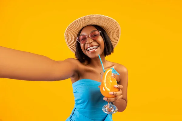 Mooie zwarte dame in trendy zomer outfit drinken verfrissende cocktail en het nemen van selfie op oranje studio achtergrond — Stockfoto