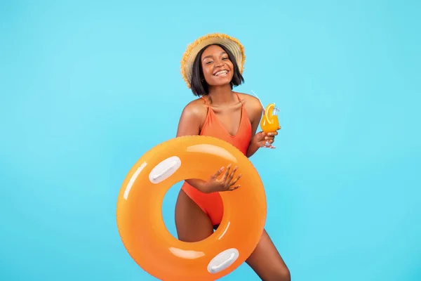 Senhora afro-americana feliz com anel inflável e saboroso coquetel tropical sorrindo para a câmera no fundo do estúdio azul — Fotografia de Stock
