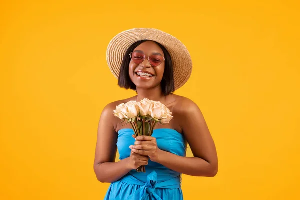 Attractive black woman in summer dress and straw hat holding bouquet of flowers on orange studio background — Stock Photo, Image