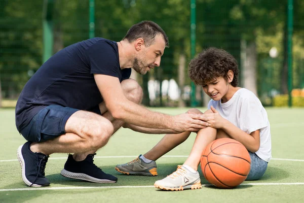 Teacher helping boy with knee trauma after playing basketball — Stock Photo, Image