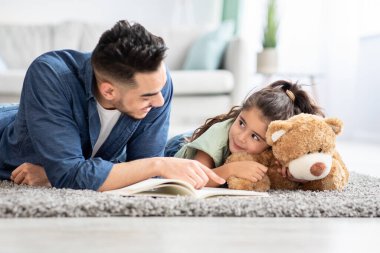 Ortadoğulu genç adam evde küçük kızıyla kitap okuyor.