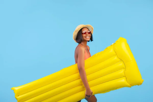 Молода чорна леді в купальнику і солом'яному капелюсі тримає жовтий надувний ліло на синьому студійному фоні — стокове фото