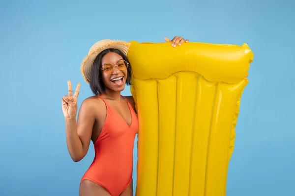 Alegre dama negra en traje de baño sosteniendo lilo amarillo y mostrando gesto de paz sobre fondo de estudio azul —  Fotos de Stock