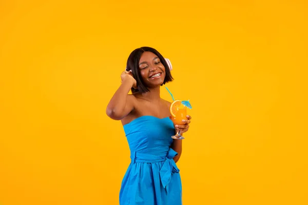 Black lady in casual outfit holding tropical summer cocktail and listening to music in headphones on orange background — Stock Photo, Image