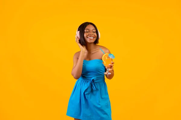 Summer party concept. Sexy black woman with tasty cocktail listening to music in headphones on orange studio background — Stock Photo, Image