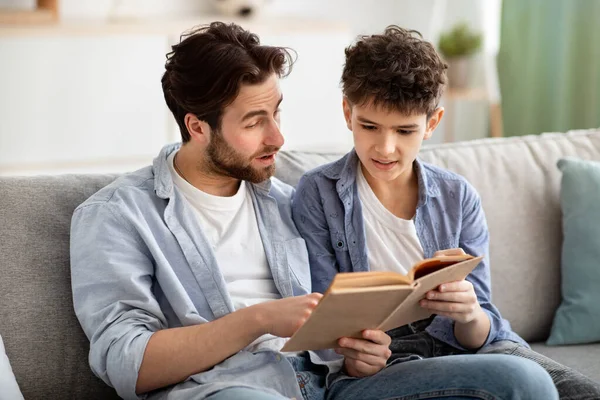 Un passe-temps familial. Père et adolescent garçon lecture livre ensemble, reposant sur canapé confortable dans le salon — Photo